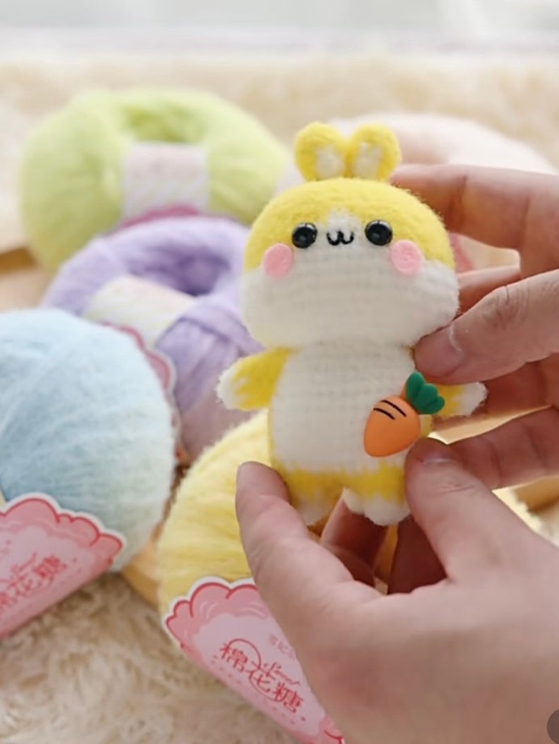 Puede incluir: Un conejito de crochet amarillo y blanco sosteniendo una peque&ntilde;a zanahoria naranja. El conejito tiene ojos negros y mejillas rosadas. El conejito est&aacute; rodeado de ovillos de lana de varios colores.