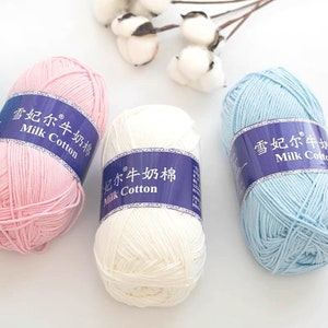 Pu&ograve; includere: Tre gomitoli di filato rosa, bianco e azzurro chiaro. Ogni gomitolo ha un'etichetta blu con il testo "雪妃尔牛奶棉 Milk Cotton" in cinese e inglese.