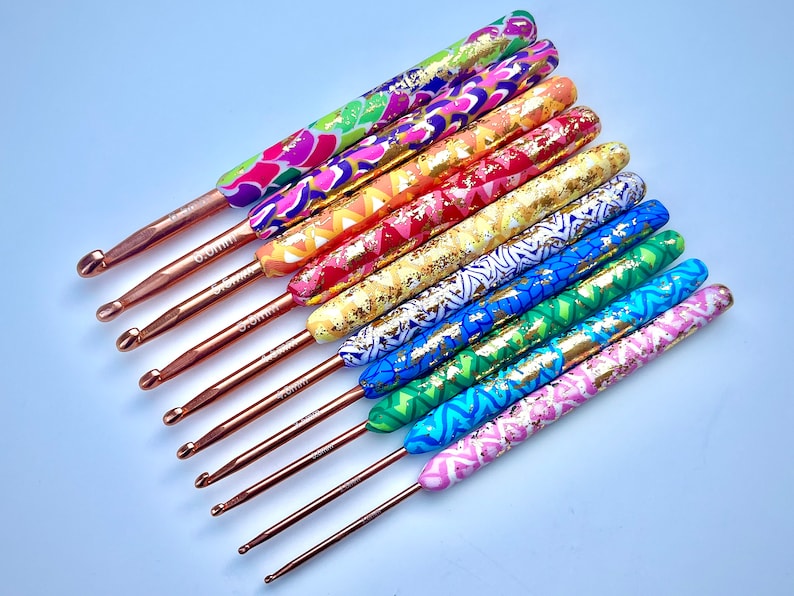 Puede incluir: Conjunto de 9 ganchillos de crochet coloridos con detalles dorados y puntas de cobre. Cada gancho tiene un patr&oacute;n y un color diferente, incluyendo rosa, azul, verde, amarillo y morado. Los ganchillos est&aacute;n dispuestos en forma de abanico, con el gancho m&aacute;s grande en la parte superior y el m&aacute;s peque&ntilde;o en la parte inferior.