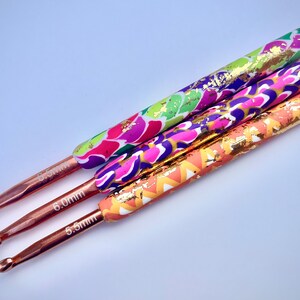 Puede incluir: Tres ganchillos de crochet coloridos con detalles de pan de oro. Los ganchillos son de 5,5 mm, 6,0 mm y 6,5 mm.