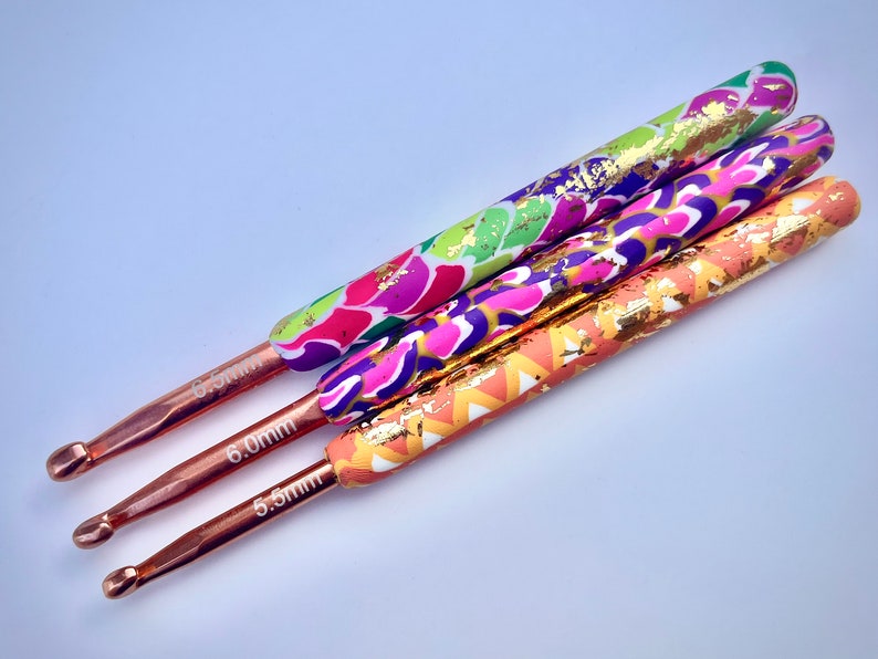 Puede incluir: Tres ganchillos de crochet coloridos con detalles de pan de oro. Los ganchillos son de 5,5 mm, 6,0 mm y 6,5 mm.