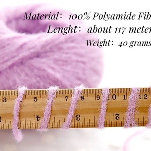 Puede incluir: Una madeja de hilo de color lila claro con una regla de madera que muestra la longitud del hilo. El hilo es 100% poliamida, de unos 117 metros de largo y pesa 40 gramos.