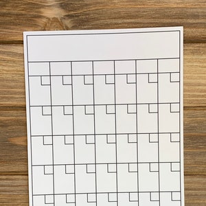 May include: A blank white monthly calendar with a grid of squares, resting on a wooden surface. The calendar has a blank header at the top and is designed for writing in dates and notes. The wood has a warm brown tone.