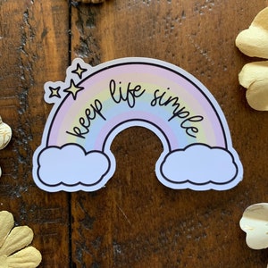 May include: A sticker featuring a pastel rainbow with the words "keep life simple" in black script. The rainbow has a white outline and is decorated with two stars and fluffy white clouds. The sticker is on a wooden surface.