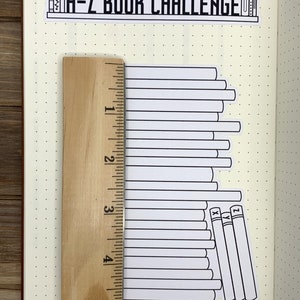 May include: A-Z Book Challenge sticker with a stack of books, a ruler, and a dotted notebook page. The sticker shows a stack of books with the letters X, Y, and Z on the spines of the last three books.