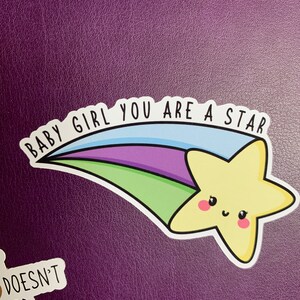 May include: A yellow star with a smiling face, on a blue, green, purple and white shooting star with the text "Baby Girl You Are A Star".