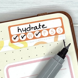 K&ouml;nnte beinhalten: Eine Nahaufnahme einer Planerseite mit einem Aufkleber, auf dem "hydrate" steht und der f&uuml;r jeden Tag der Woche Kontrollk&auml;stchen hat. Der Aufkleber ist orange und wei&szlig; mit schwarzen H&auml;kchen. Ein grauer Stift liegt auf der Seite.