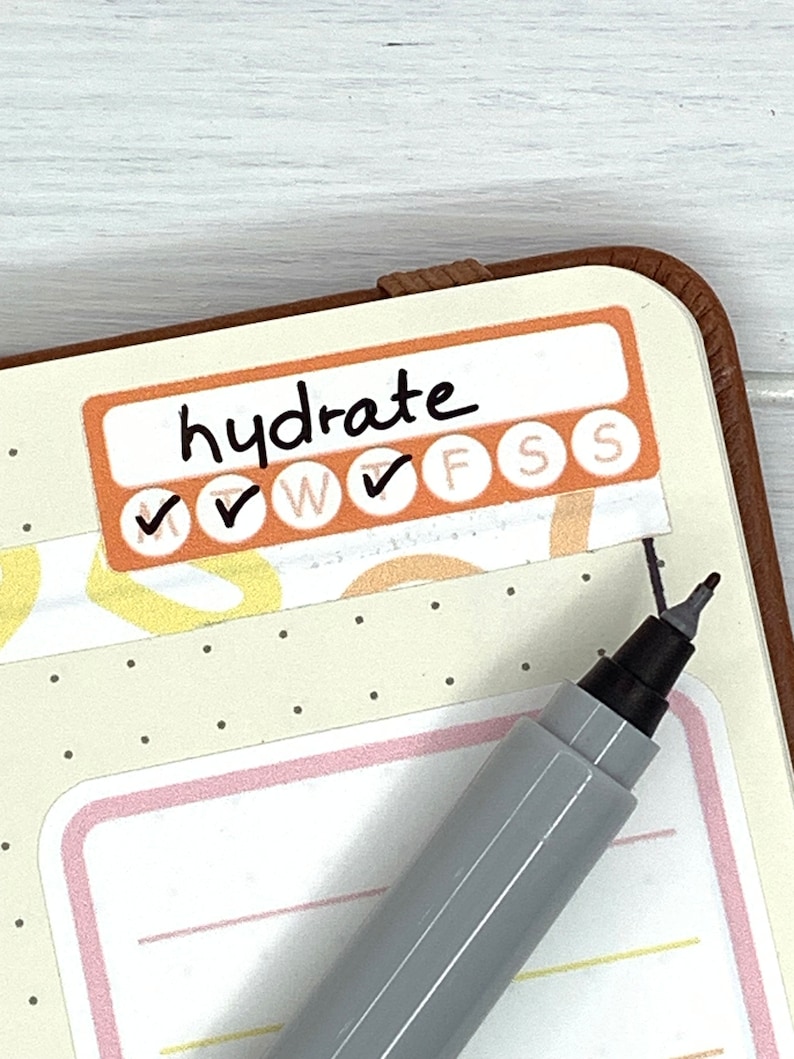 K&ouml;nnte beinhalten: Eine Nahaufnahme einer Planerseite mit einem Aufkleber, auf dem "hydrate" steht und der f&uuml;r jeden Tag der Woche Kontrollk&auml;stchen hat. Der Aufkleber ist orange und wei&szlig; mit schwarzen H&auml;kchen. Ein grauer Stift liegt auf der Seite.