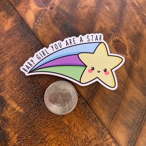 May include: A yellow star with a smiling face, with a rainbow tail and the text "Baby Girl You Are A Star" on a wooden background.