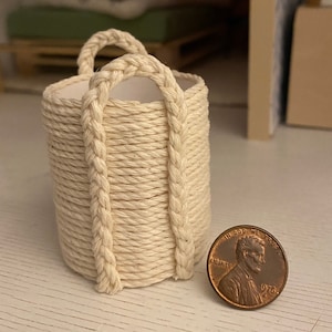 May include: A small, cream-colored rope basket with two handles. The basket is made of woven rope and has a natural, rustic look. A penny is placed next to the basket for scale.