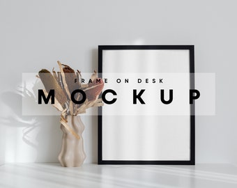 May include: A black picture frame mockup on a white desk with a vase of dried flowers. The text "FRAME ON DESK MOCKUP" is visible in the image.