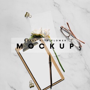May include: A white card with a flower on a marble surface. A clipboard with a blank white sheet of paper and a pen is next to the card. The text "CARDS WITH FLOWER MOCKUP" is written on the image.
