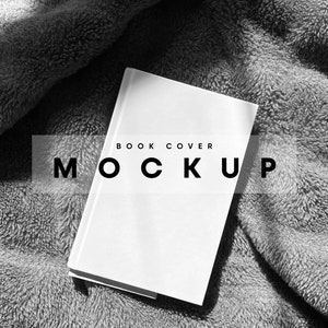 Op de afbeelding: Een witte boekomslag mockup op een grijs, pluche deken. De tekst "BOOK COVER MOCKUP" is op de omslag gedrukt.