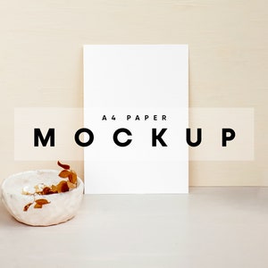 May include: A white A4 paper mockup with the text "A4 PAPER MOCKUP" on a light beige background. A small white ceramic bowl with dried brown leaves sits in the foreground.