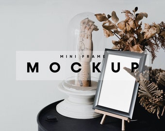 May include: A black picture frame with a white mat and a white background is on a wooden easel. The frame is on a black surface with a glass dome and a hand sculpture inside. The text "MINI FRAME MOCKUP" is on a white rectangle in the background.