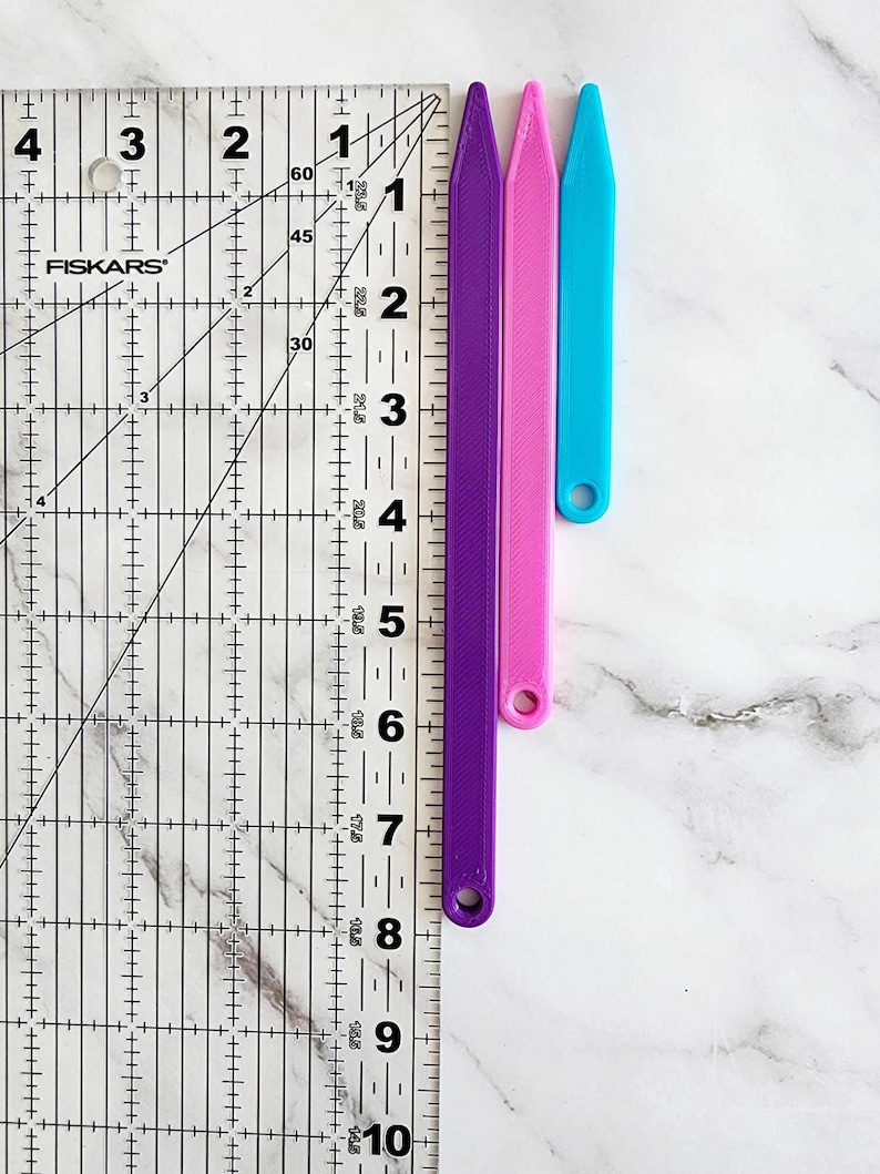 May include: Three plastic tools for crafting, measuring 7 inches in length. The tools are purple, pink, and blue. The tools are shown against a white marble background.