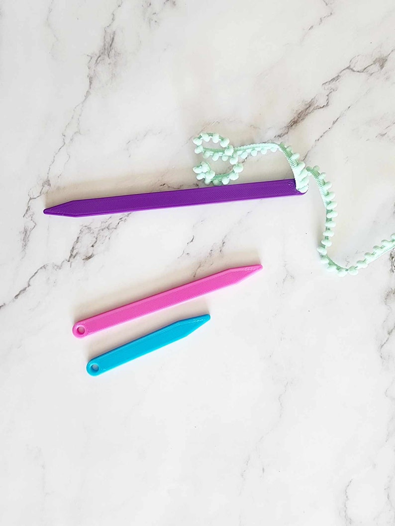 May include: Three plastic tools for crafting, one purple, one pink, and one blue. The tools are on a white marble surface.