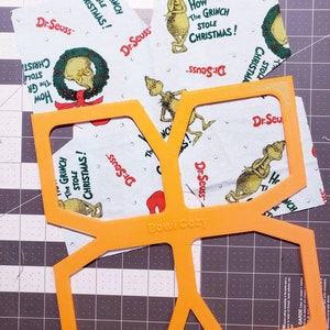 May include: Orange plastic template for sewing a bowl cosy. The template is labelled "Bowl Cozy" and has a design for a hexagon-shaped cosy. The template is on top of a cutting mat with a grid pattern. There are several pieces of fabric with a Grinch design on top of the cutting mat.