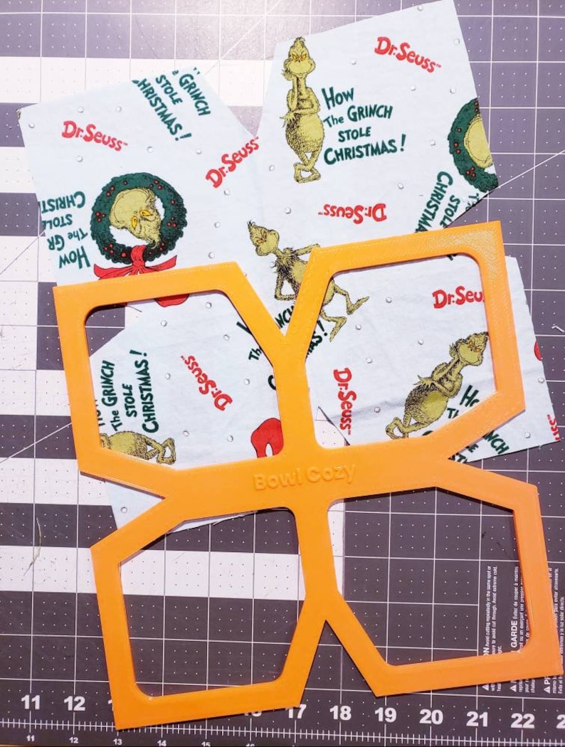 May include: Orange plastic template for sewing a bowl cosy. The template is labelled "Bowl Cozy" and has a design for a hexagon-shaped cosy. The template is on top of a cutting mat with a grid pattern. There are several pieces of fabric with a Grinch design on top of the cutting mat.