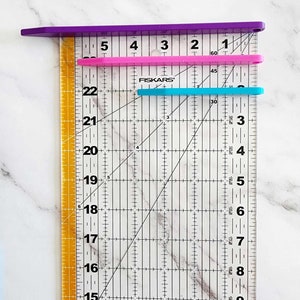 May include: A clear ruler with black markings and a diagonal line. The ruler has numbers from 14 to 22 on the left side and numbers from 1 to 60 on the top. There are three rulers on top of the clear ruler, one purple, one pink, and one blue.
