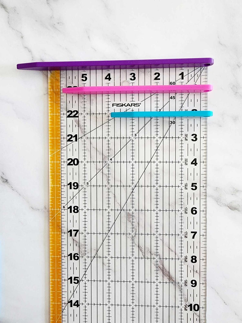 May include: A clear ruler with black markings and a diagonal line. The ruler has numbers from 14 to 22 on the left side and numbers from 1 to 60 on the top. There are three rulers on top of the clear ruler, one purple, one pink, and one blue.