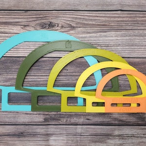 May include: Four plastic semi-circle shaped stencils in different sizes. The largest stencil is light blue, the next largest is green, the next is yellow, and the smallest is orange.