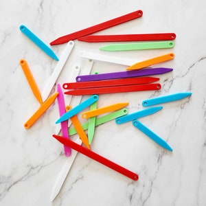 May include: A collection of colorful plastic knitting needles, arranged in a random pattern on a white marble surface.