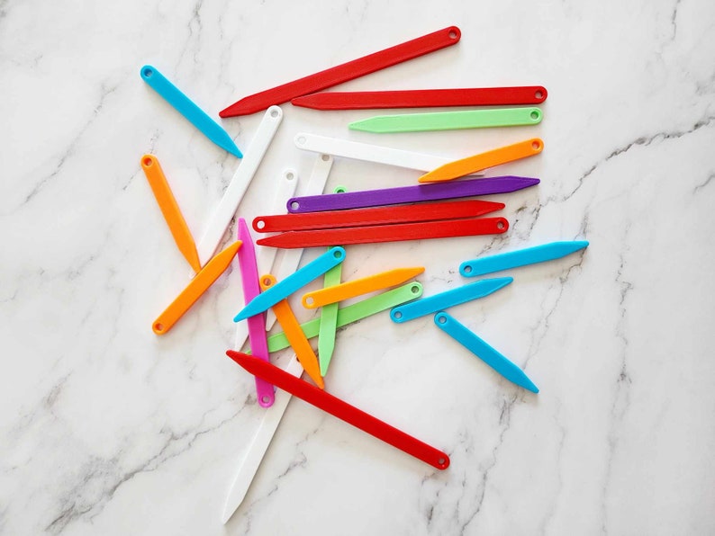 May include: A collection of colorful plastic knitting needles, arranged in a random pattern on a white marble surface.