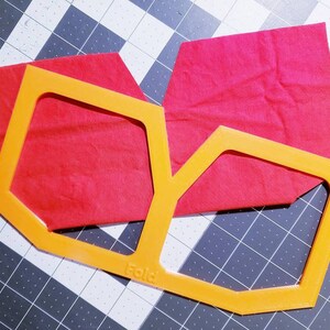 May include: Orange plastic template for cutting fabric, with a red fabric piece on top. The template has the word "Fold" printed on it.