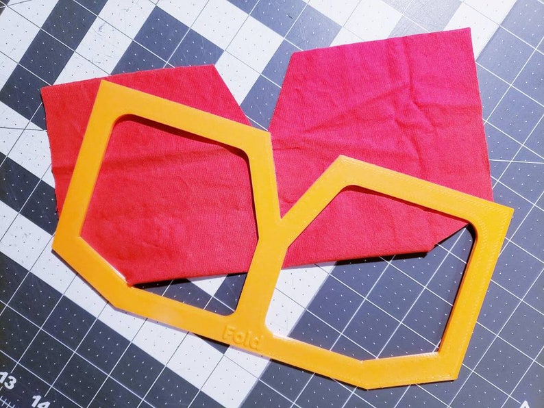 May include: Orange plastic template for cutting fabric, with a red fabric piece on top. The template has the word "Fold" printed on it.