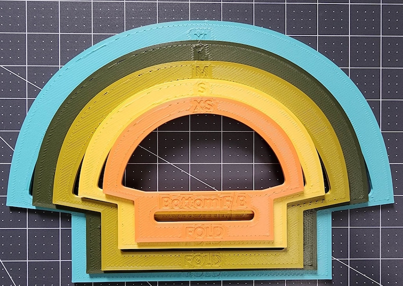 May include: Five rainbow-colored fabric cutting templates labeled with sizes XS, S, M, and L. The templates are shaped like half circles with a fold line marked on each. The bottom template has the words "Bottom 1/2 Fold" printed on it.