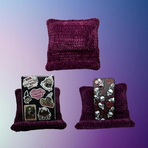 May include: Two purple plush phone stands, one with a phone case featuring a skull and rose design, the other with a phone case decorated with stickers that read "A book a day keeps reality away" and "Rather be reading".