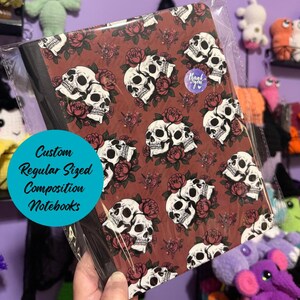 Peut inclure: Un cahier de composition avec un motif répété de crânes blancs et de roses rouges sur un fond marron. Le cahier est dans une pochette en plastique transparent. Le texte sur le cahier indique "Custom Regular Sized Composition Notebooks".