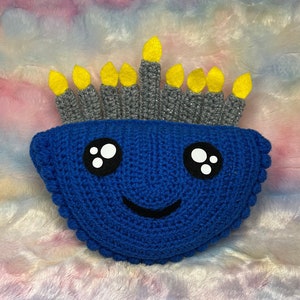 May include: A blue crocheted pillow shaped like a birthday cake with yellow candles and a smiling face.