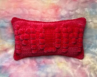 May include: A bright pink crocheted pillow with a textured surface. The pillow is on a fluffy, multi-colored background.