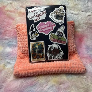 Op de afbeelding: Een zwarte telefoonhoes met verschillende stickers, waaronder een skelet dat een boek leest, een kopje thee en boeken, en de tekst "Rather be reading".