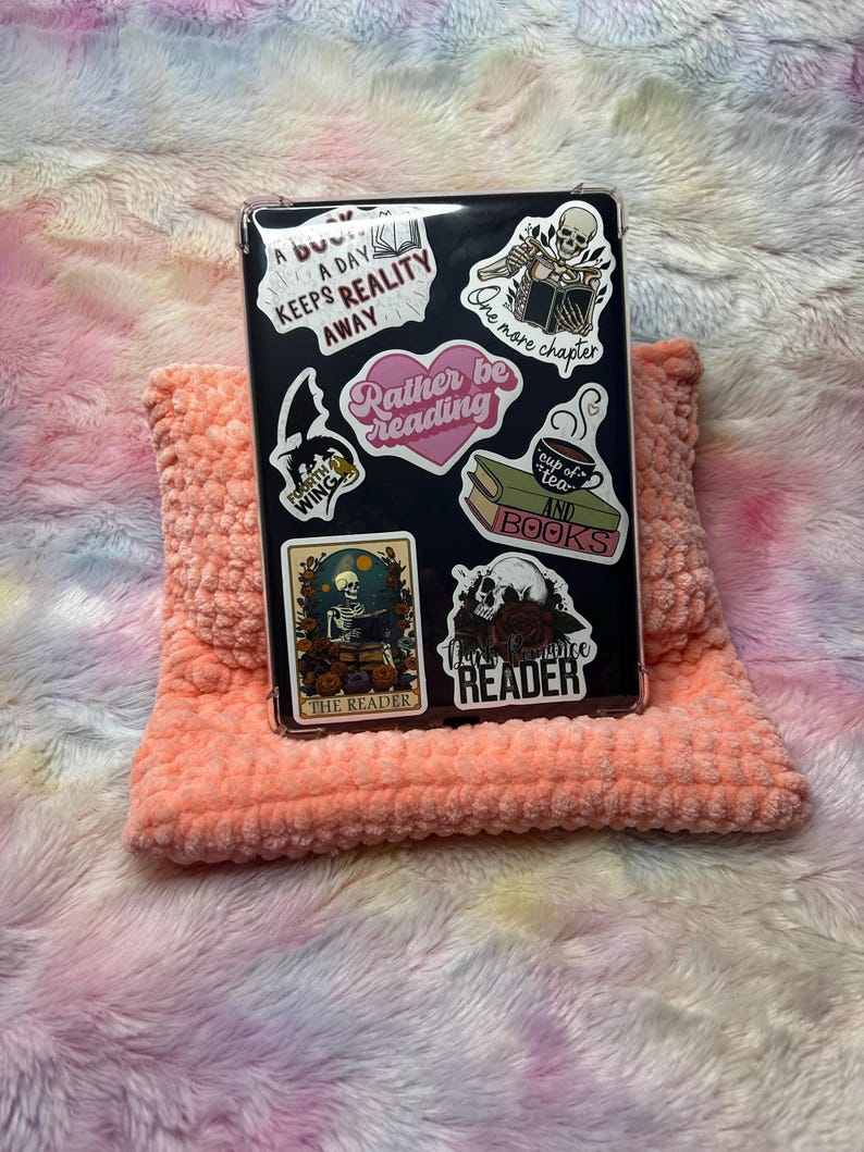Op de afbeelding: Een zwarte telefoonhoes met verschillende stickers, waaronder een skelet dat een boek leest, een kopje thee en boeken, en de tekst "Rather be reading".