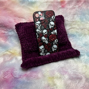Op de afbeelding: Een zwarte telefoonhoes met een schedel- en rozenontwerp op een paars kussen. De telefoonhoes ligt op een wit en roze pluche oppervlak.
