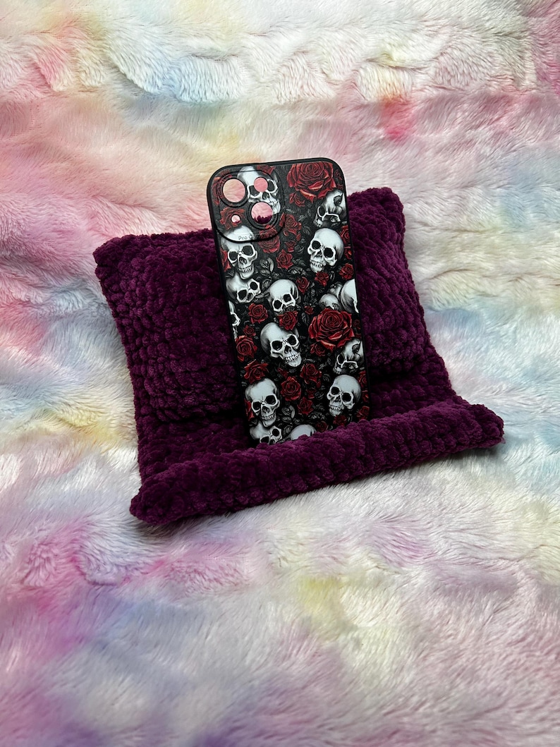 Op de afbeelding: Een zwarte telefoonhoes met een schedel- en rozenontwerp op een paars kussen. De telefoonhoes ligt op een wit en roze pluche oppervlak.