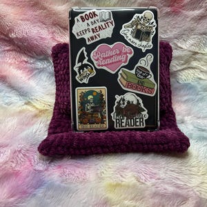 Op de afbeelding: Een transparante telefoonhoes met verschillende kleurrijke stickers met citaten over lezen. De stickers bevatten "Een boek per dag houdt de realiteit weg", "Liever lezen", "Nog een hoofdstuk", "Kop thee en boeken", "De lezer" en "De vraatzuchtige lezer".