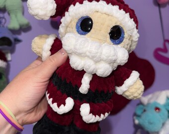 May include: A handmade crochet Santa Claus doll with a red velvet suit, black boots, and a white beard and trim. The doll has large, glittery blue eyes and a matching red hat with a white pom-pom. Other crochet toys are visible in the background.