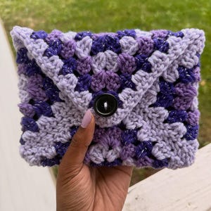 May include: A handmade crochet clutch in shades of purple, lavender, and silver. The clutch features a granny square design and a black button closure. The folded flap secures the contents. The clutch is held in a hand.
