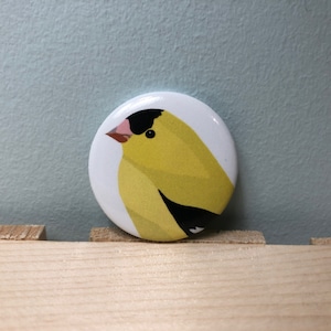 Pode incluir: Um botão redondo com uma ilustração de um pássaro amarelo e preto. O pássaro está virado para a esquerda e tem uma cabeça preta e um corpo amarelo com uma asa preta.