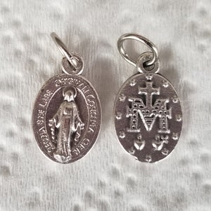 Peut inclure: Deux breloques en m&eacute;tal argent&eacute; avec des images religieuses. Un charme repr&eacute;sente la Vierge Marie avec le texte "ORIGINAL SIN LARE" et "CONCEPCIA ORA". L'autre charme repr&eacute;sente la lettre "M" avec une croix au-dessus et deux c&oelig;urs en dessous.