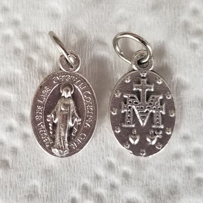 Peut inclure: Deux breloques en m&eacute;tal argent&eacute; avec des images religieuses. Un charme repr&eacute;sente la Vierge Marie avec le texte "ORIGINAL SIN LARE" et "CONCEPCIA ORA". L'autre charme repr&eacute;sente la lettre "M" avec une croix au-dessus et deux c&oelig;urs en dessous.