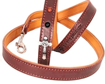 May include: A brown leather dog lead with a cross charm and the words "Live on the Journey" embossed on the strap. The lead has a silver clip and orange stitching.