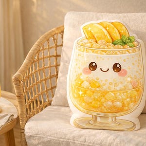 Puede incluir: Un cojín con forma de vaso de té de burbujas de mango de dibujos animados, con una cara sonriente, rodajas de mango y hojas de menta. El cojín está sobre un cojín blanco, junto a un vaso de té de burbujas de mango.