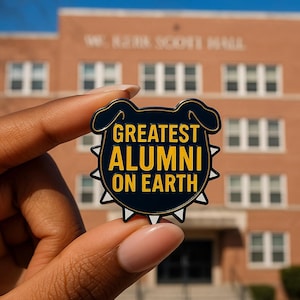 May include: A navy blue enamel pin with the words "GREATEST ALUMNI ON EARTH" in yellow. The pin has a black border and white accents. The pin is held in front of a brick building.