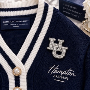 May include: A navy blue knit cardigan with white striped trim, featuring a Hampton University label, a rhinestone-encrusted HU logo pin, and white pearl-style buttons. The text Hampton Alumni is embroidered in white script on the chest.