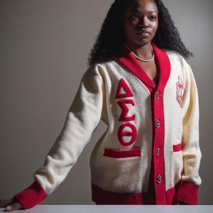 May include: Cream-coloured cardigan with red trim, displaying the Greek letters ΔΣΘ and the numbers 1913 in red. The cardigan has a V-neck, pockets, and a crest on the right side. A pearl necklace is also visible.